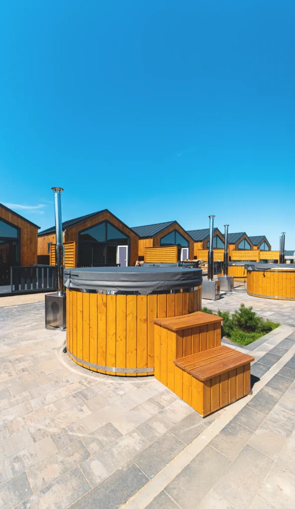 Drewniana balia z schodkami oraz piecem i kominem. Wybrukowane podłoże. W tle domki oraz błękitne niebo.