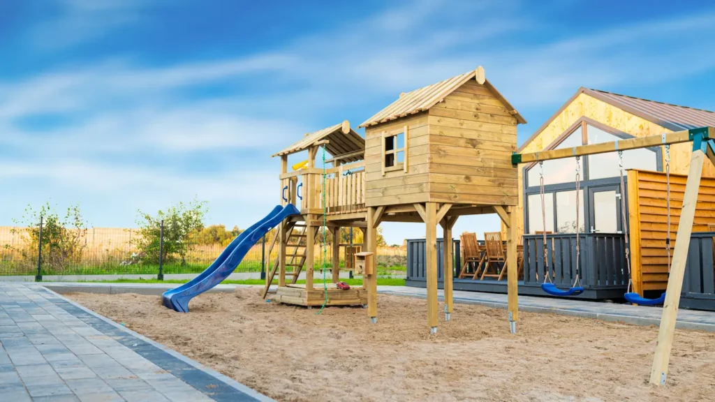 Wykonany z drewna plac zabaw dla dzieci. Niebieska zjeżdżalnia oraz zielona lina do wspinania. Dwie wieże oraz huśtawki. Piasek na podłożu.