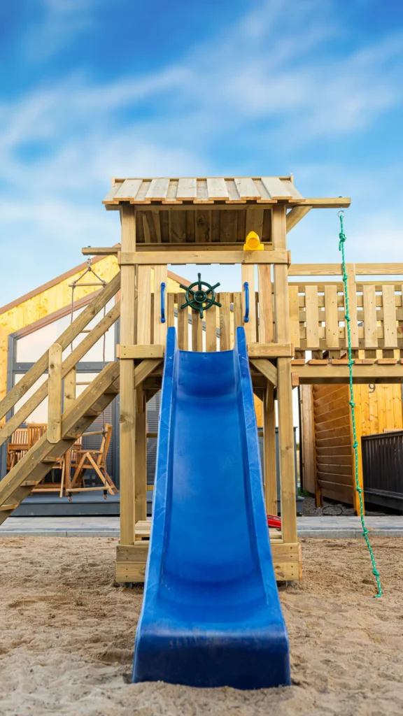 Wykonany z drewna plac zabaw dla dzieci. Niebieska zjeżdżalnia oraz zielona lina do wspinania. Piasek na podłożu.