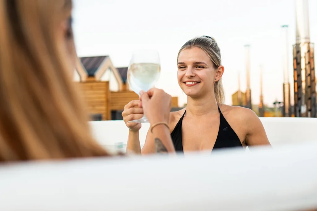 Uśmiechnięte kobiety w balii, stukające się kieliszkami z napojem. W tle domki.